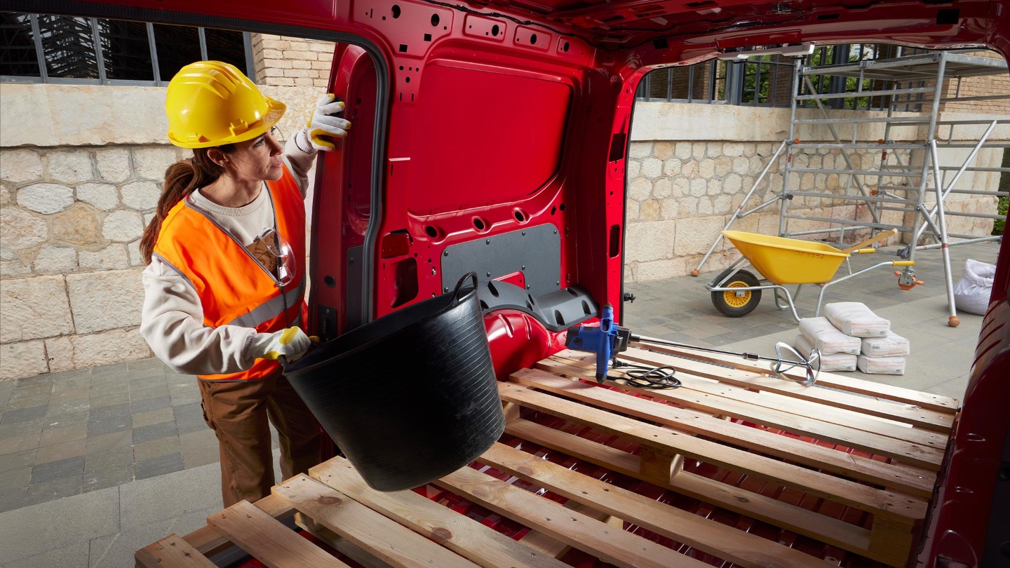 A woman places materials into the loadspace of All-New Ford Transit Connect in Maple Red with the side and back doors open, on a building site. 